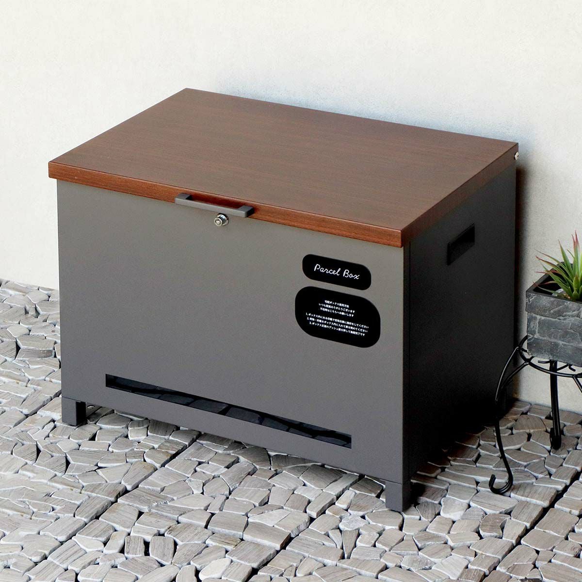 ダークグレーの宅配ボックスの設置イメージ。白い壁と石畳に置かれている。