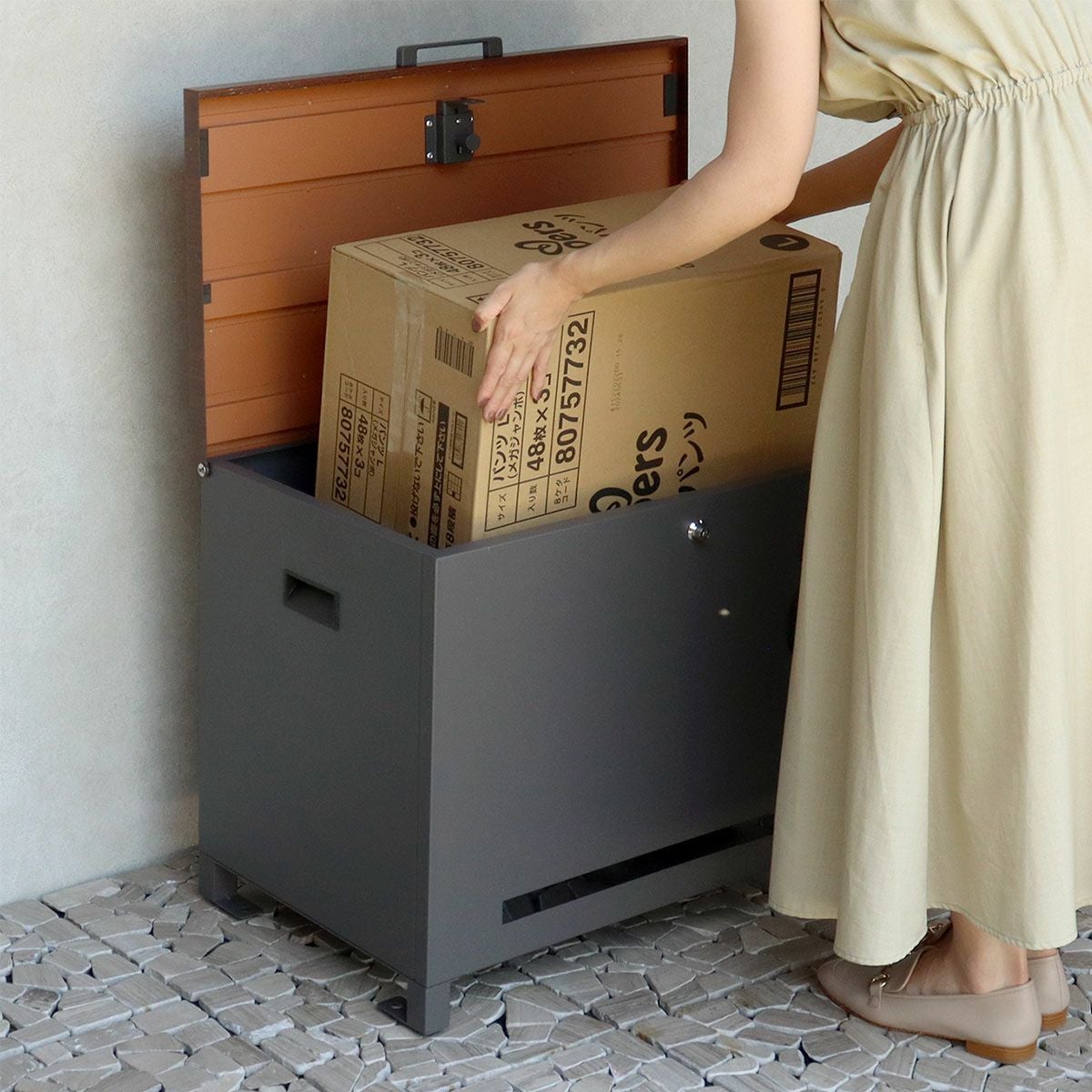 ダークグレーの宅配ボックスの設置イメージ。ワンピースを着た人が宅配ボックスから段ボールの荷物を取り出している。
