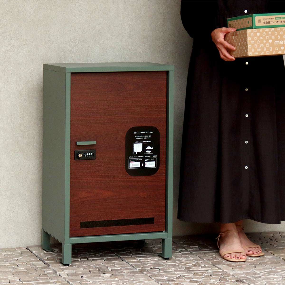 フォレストグリーンの宅配ボックスの設置イメージ。黒いワンピースを着た人が段ボールの荷物を持って側に立っている。