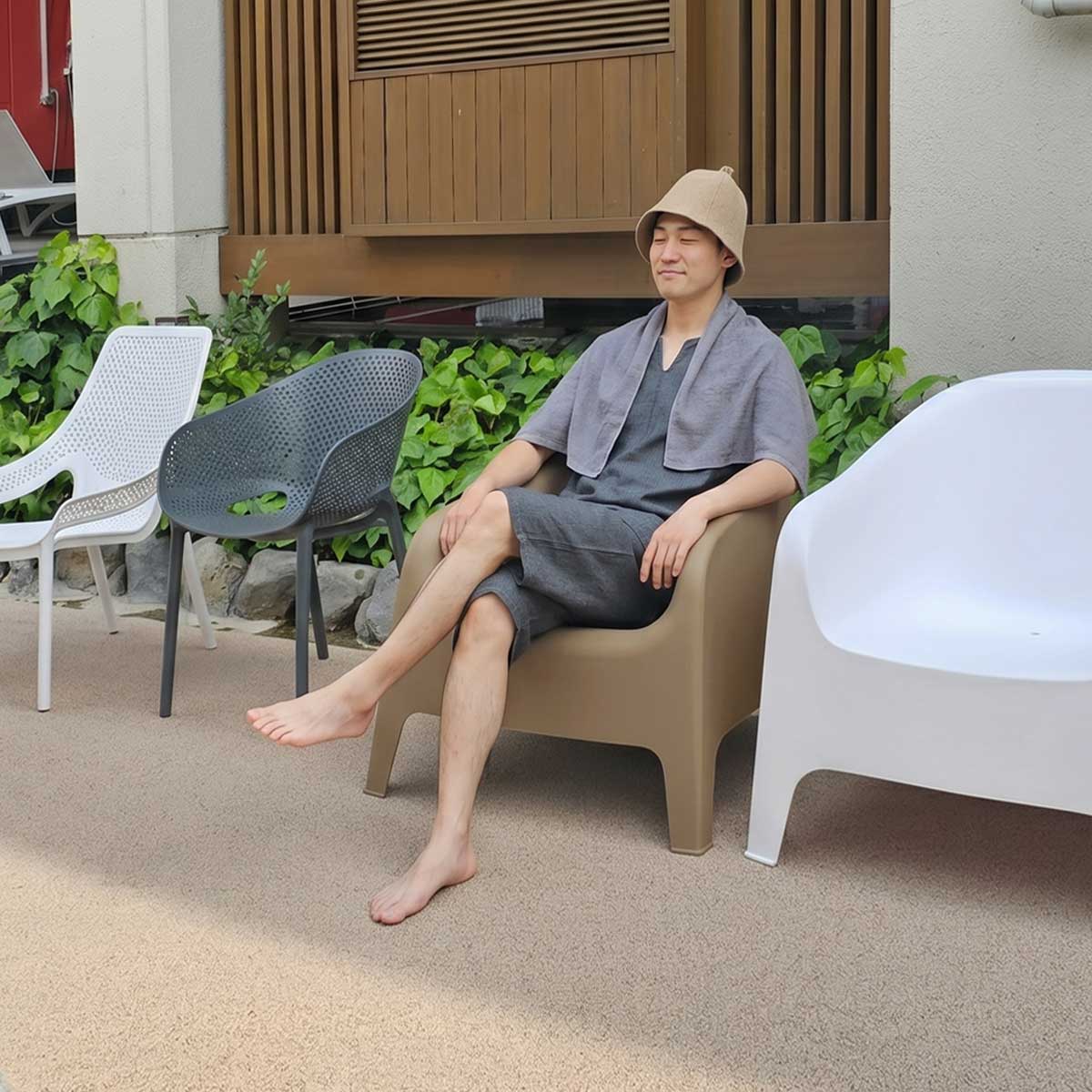 サウナウエアを着た男性が外気浴スペースのアルタス ローチェアに座り、脚を組んでゆったりとくつろいでいる設置イメージ画像です。